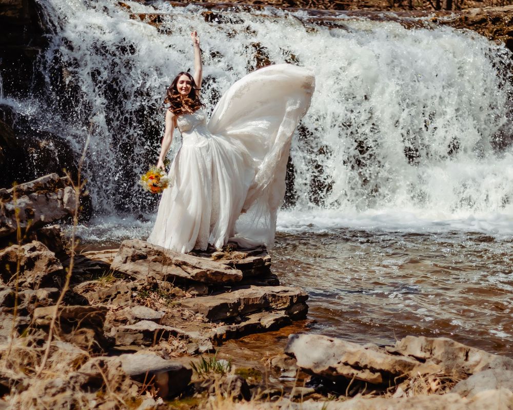 A flowing white wedding dress billows in the wind as a person poses by a rushing waterfall.
