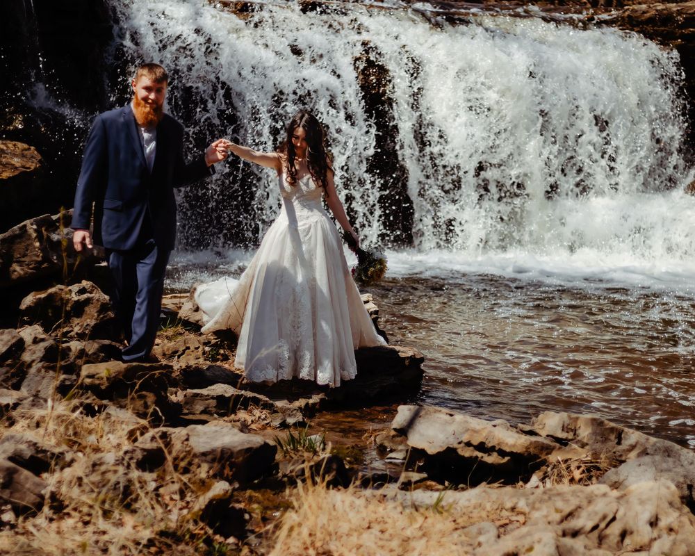 Couple shares intimate moment on rocky outcrop with waterfall rushing behind them.
