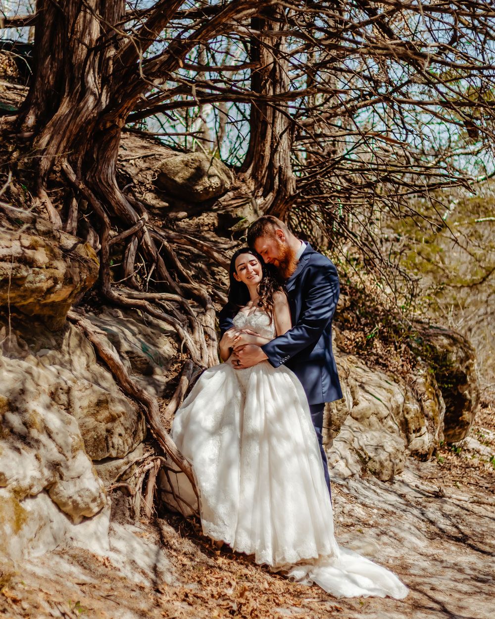 A romantic wedding photo captured under a gnarled tree on a rocky hillside with dappled sunlight.