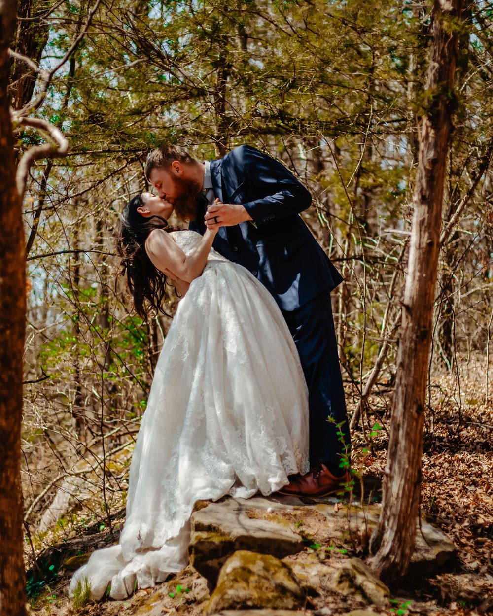 An intimate wedding moment captured on rocky terrain in a sunlit forest.