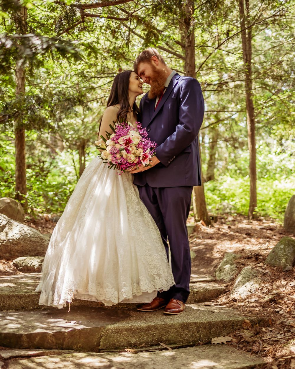 A couple embraces on stone steps in a wooded area with a pink bouquet.