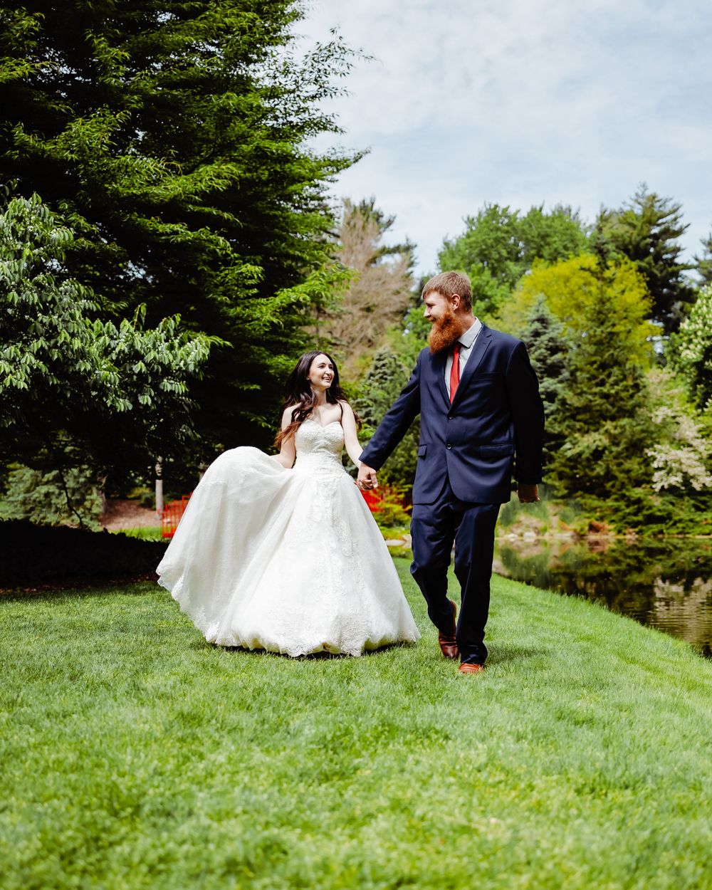 Newlyweds walking together on grassy hill surrounded by evergreen trees.