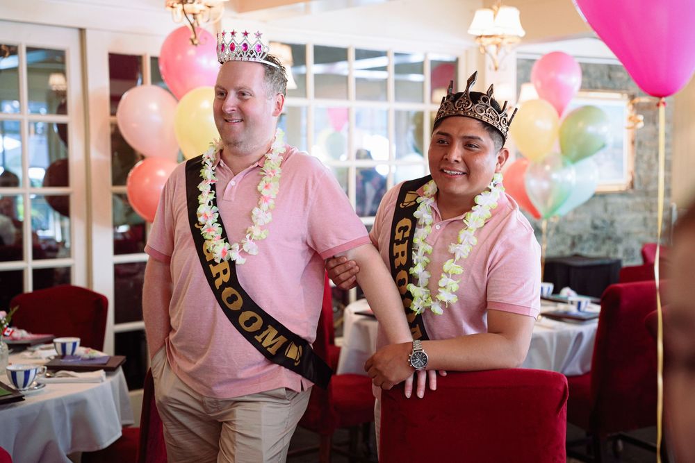 Two grooms wearing crowns, leis, and sashes celebrate at a colorful balloon-filled wedding reception venue. Toronto GTA