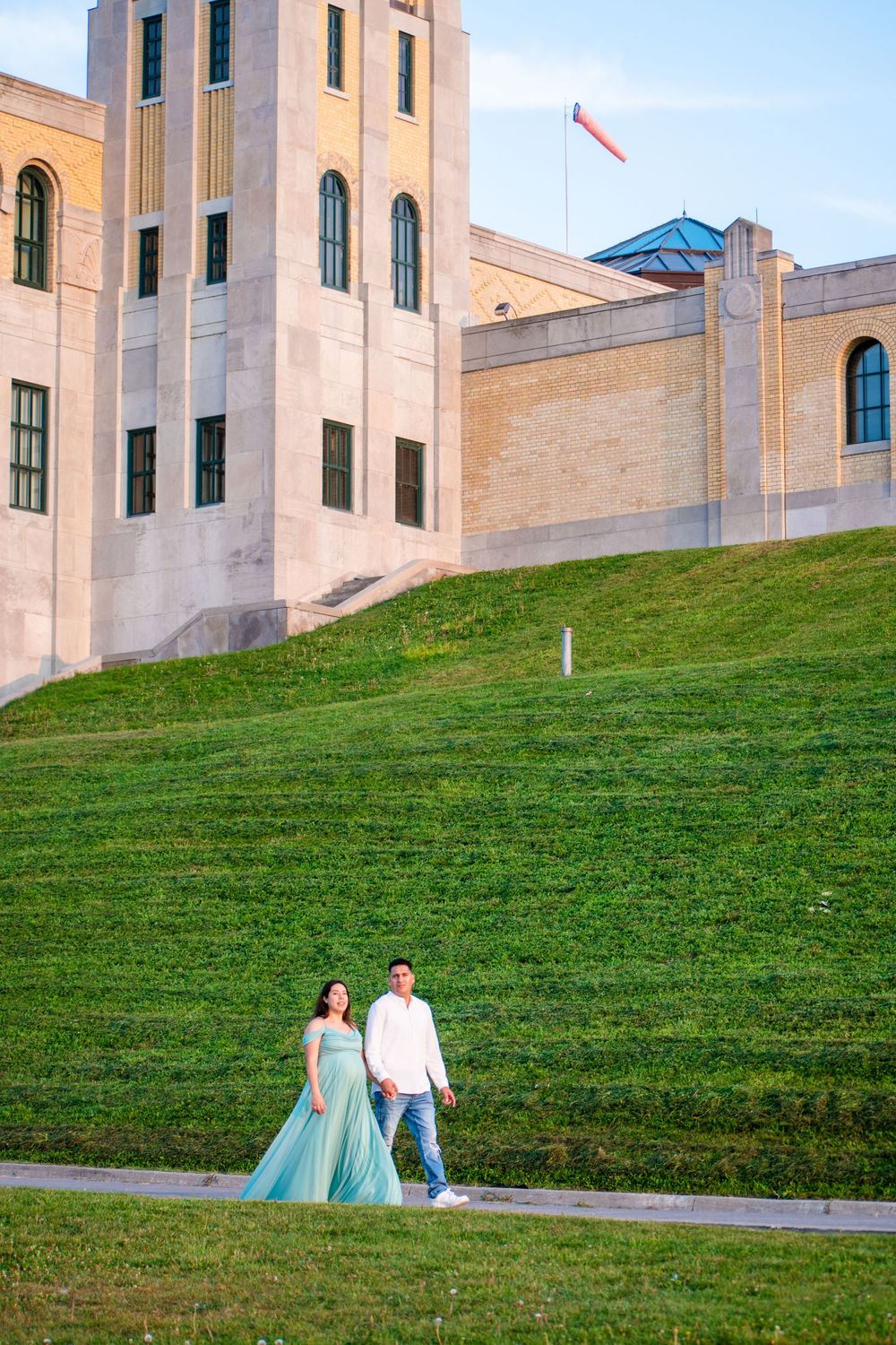 Pregnant couple in teal gown walks near grand building on green hill at golden hour. Toronto GTA