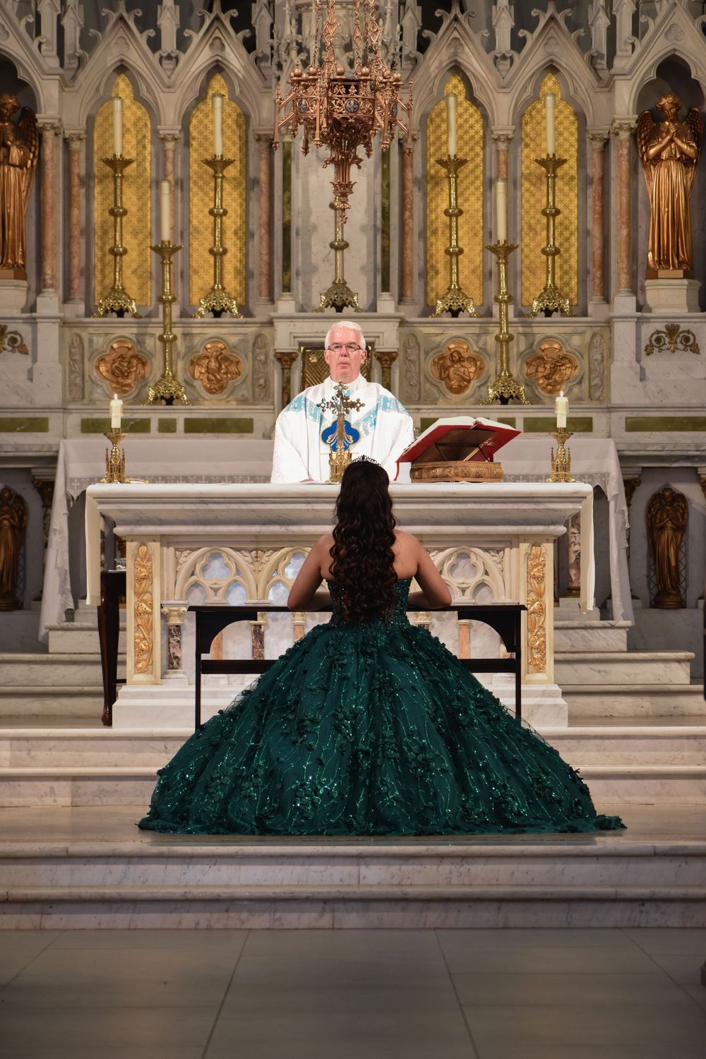Young woman in green quinceañera gown kneels before priest at ornate church altar during religious ceremony. Toronto GTA