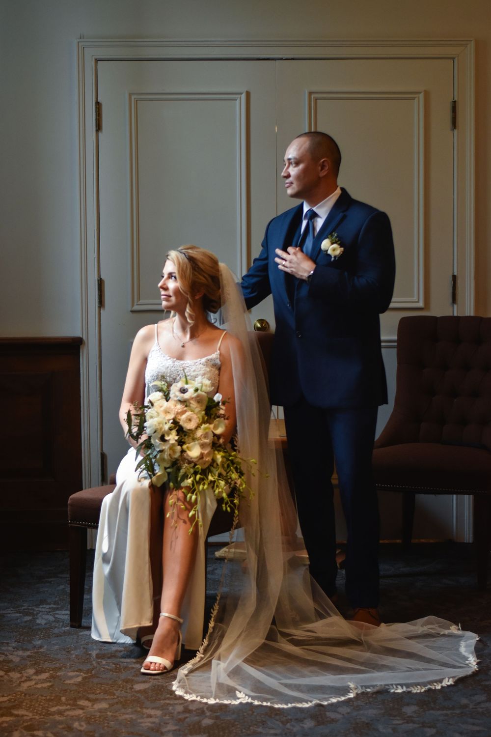 Bride seated in white gown holding floral bouquet with groom in navy suit standing beside her in elegant room. Toronto GTA