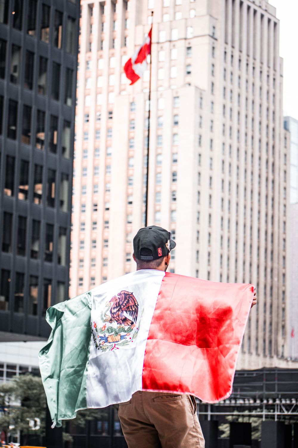 Man draped in Mexican flag stands in urban setting with Canadian flag on flagpole behind him. Toronto GTA