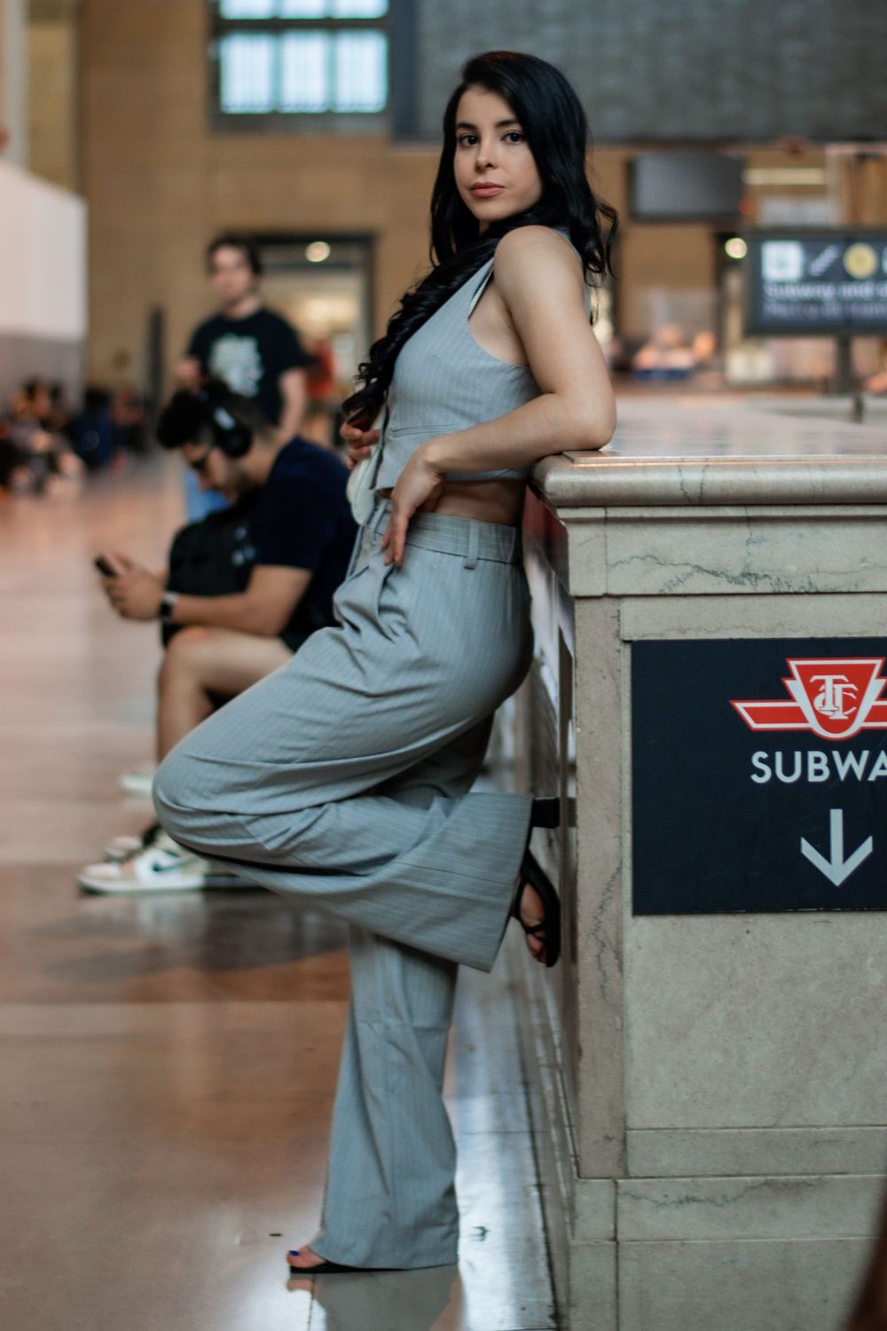 Young woman in grey outfit leans against marble pillar near subway sign in transit station. Toronto GTA
