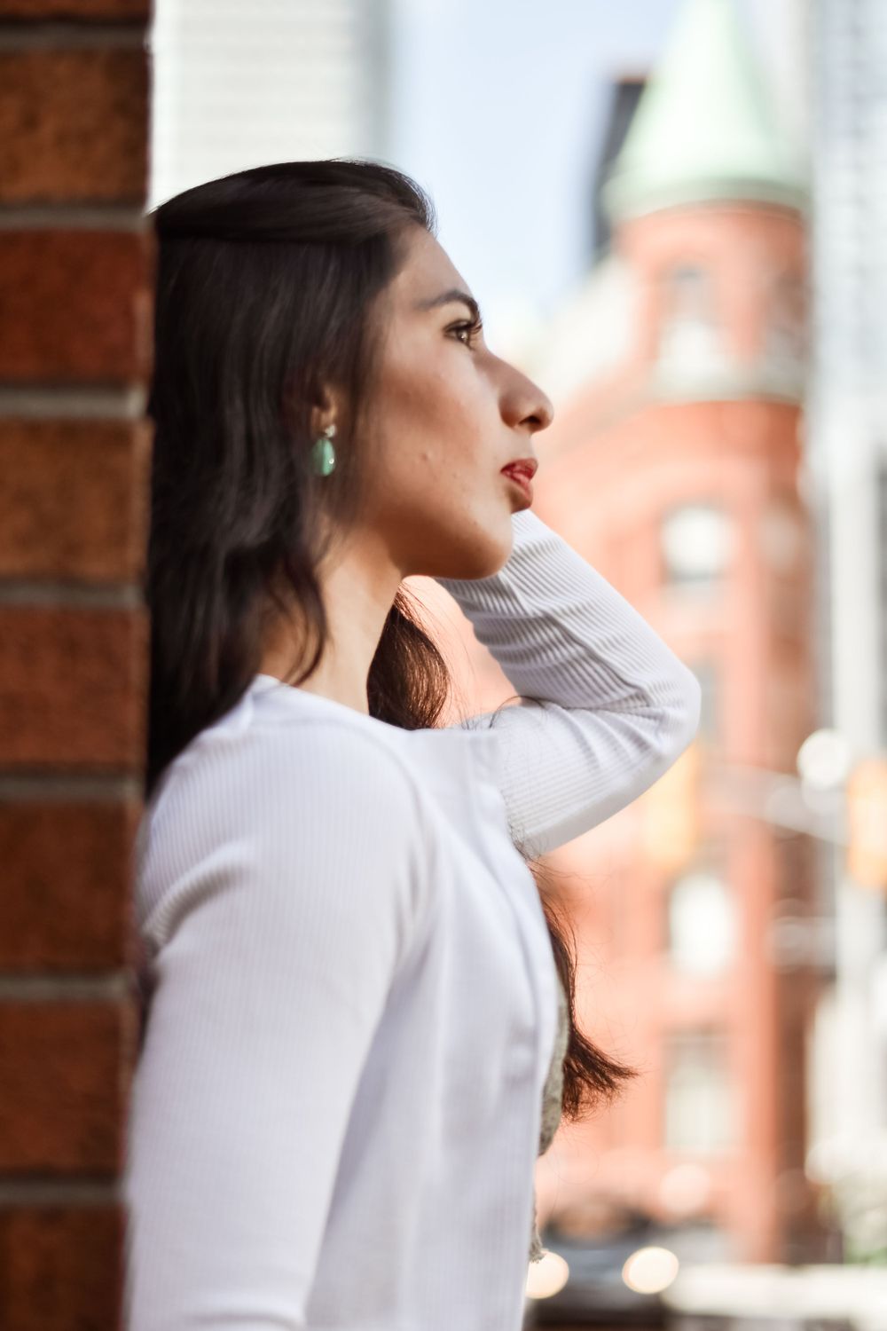 Dark-haired woman in white top leans against brick wall gazing at historic city building. Toronto GTA
