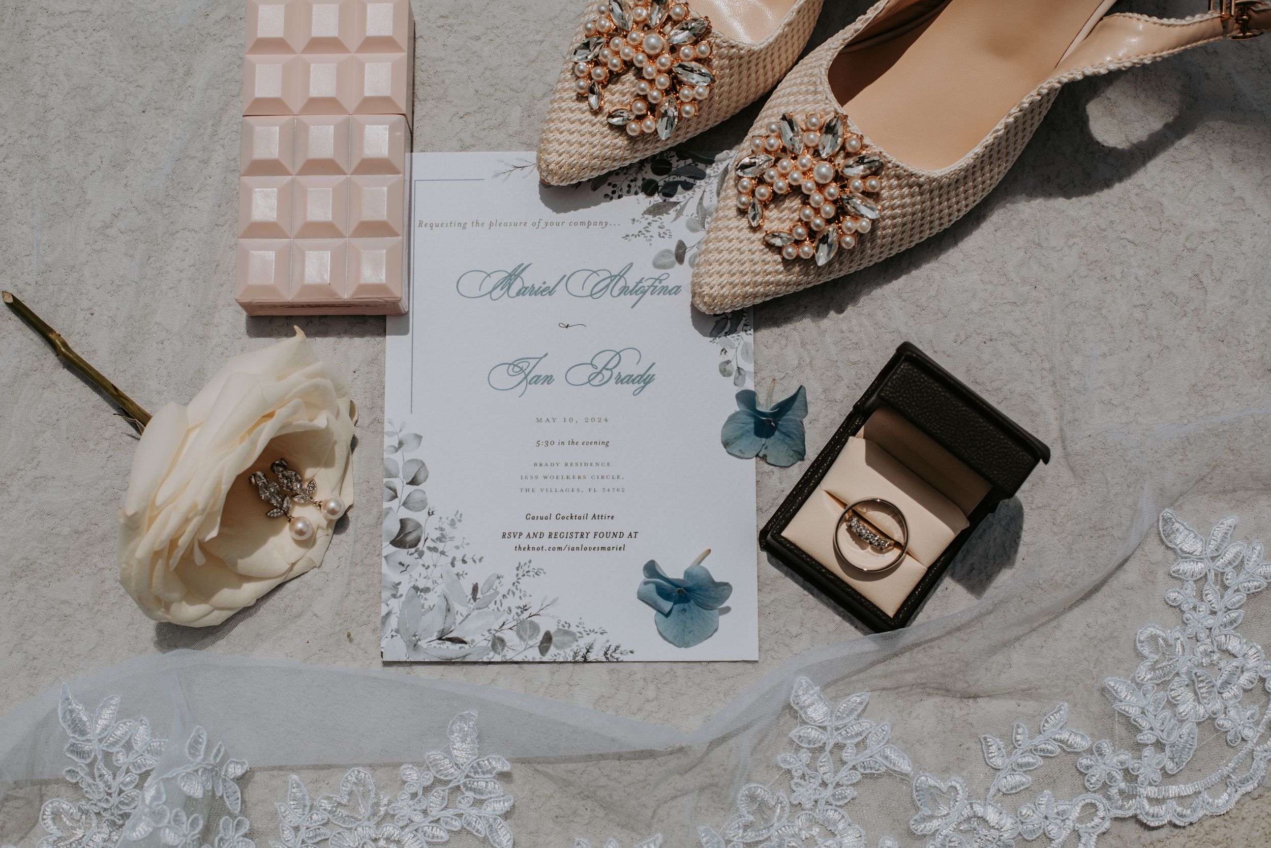 Wedding details including invitation, rings, studded flats and lace laid out on gray fabric.