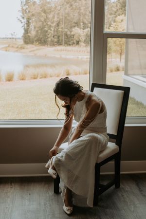 Person in wedding dress adjusting their shoe while seated by a window.