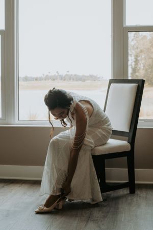 Person in wedding dress adjusting shoe while seated by window.