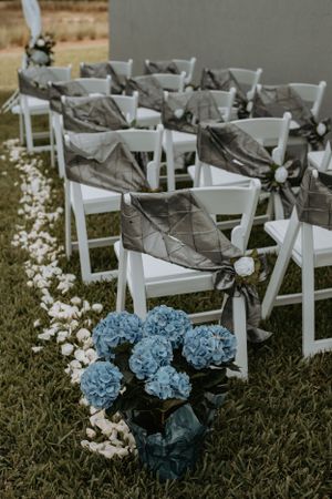White wedding ceremony chairs decorated with gray sashes and blue hydrangea floral arrangements on grass.