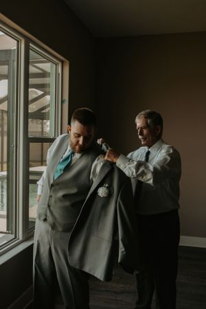 Groomsmen in gray suits prepare by a window in dramatic lighting.