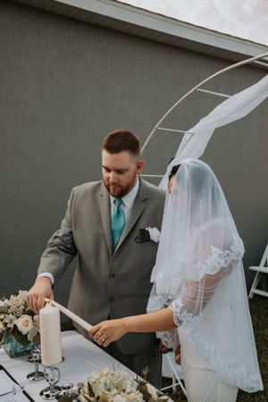 A couple performs unity candle ceremony at outdoor wedding under white arch.