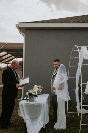 Outdoor wedding ceremony with white arch and floral decorations against grey building.