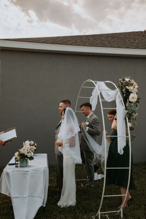 Wedding party gathered under decorative white arch during outdoor ceremony.