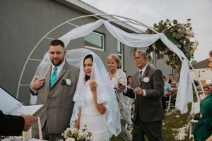 Wedding ceremony participants gather under white decorated arch during outdoor service.