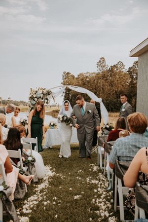 Outdoor wedding ceremony in a field with white chairs and wildflowers as guests gather under cloudy skies.