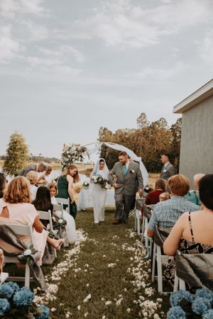 Wedding guests seated in white chairs on a grassy field with wildflowers during an outdoor ceremony.