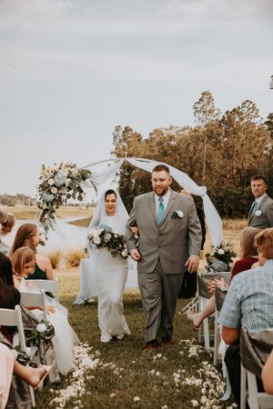 Wedding party poses together on a grassy field with white chairs and autumn trees in the background.