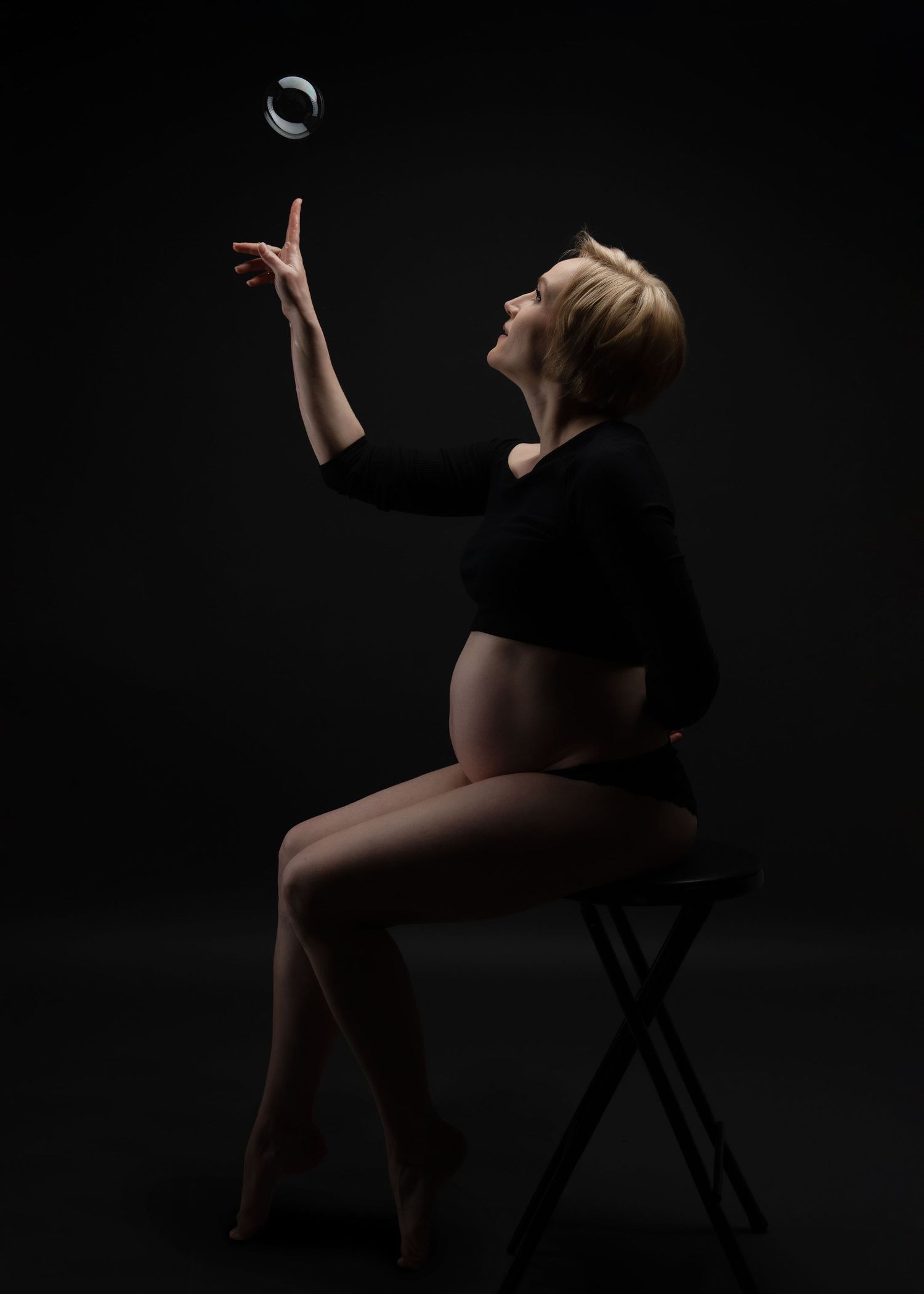 maternity photo shoot in studio, a pregnant woman looks up at a floating bubble with a finger posed to pop it