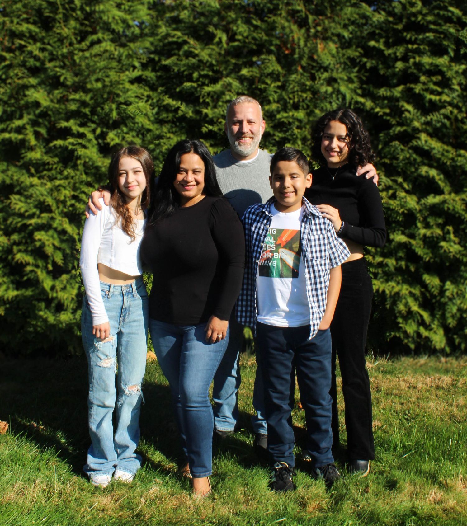 Family of five posing outdoors against a green hedge background.
