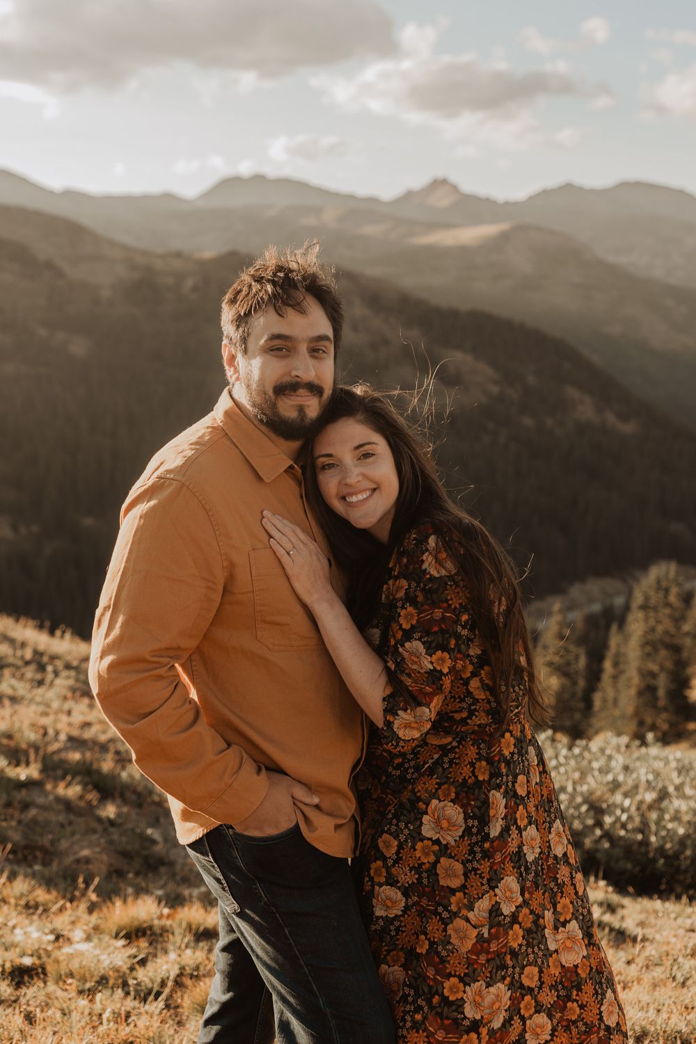 Couple embraces during golden hour on mountainside with panoramic mountain views in background.