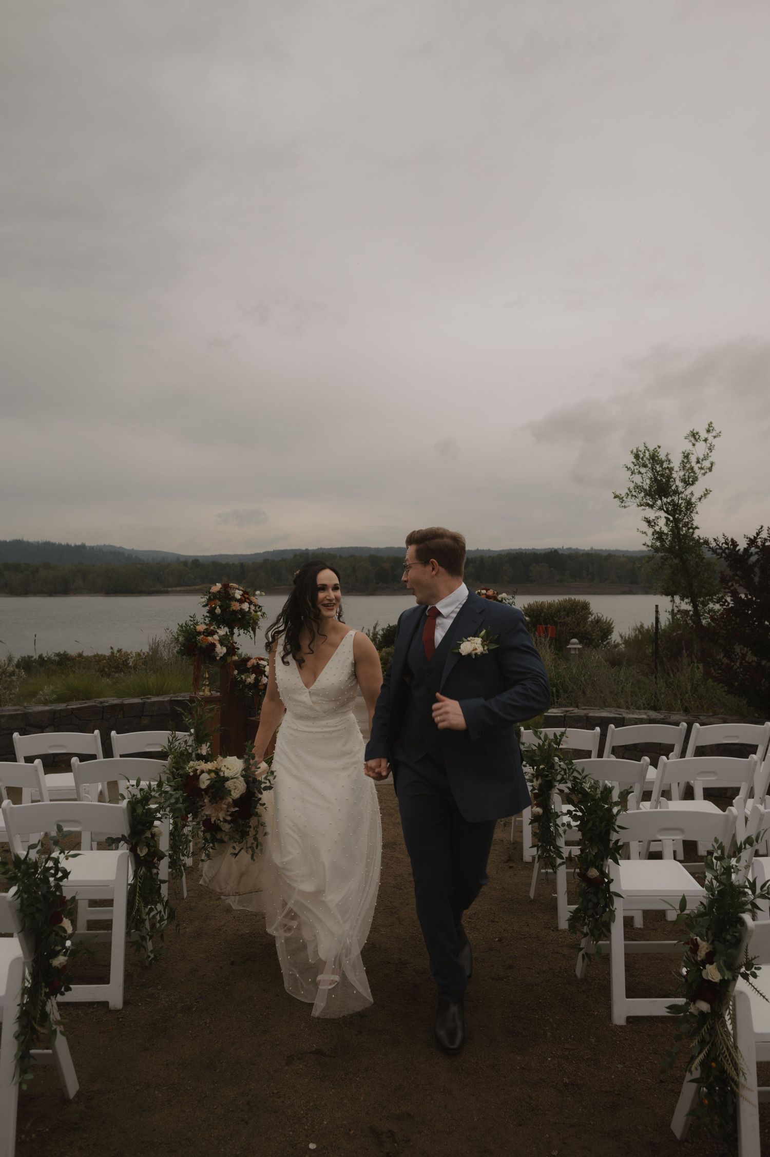 Couple walks down aisle between white chairs with floral arch and lake view.
