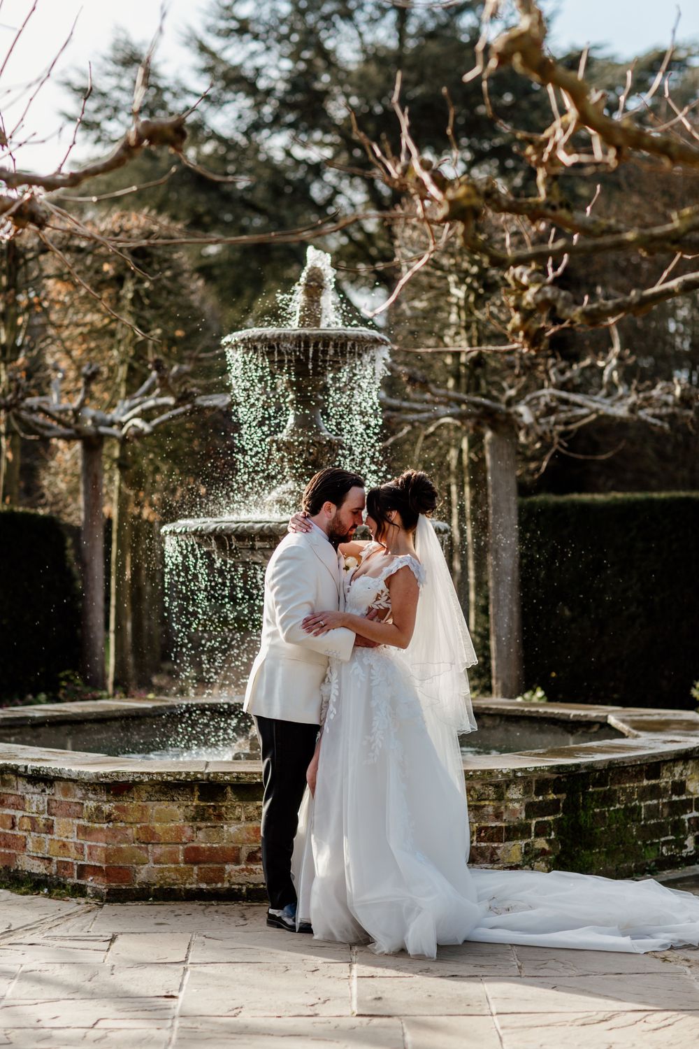 A romantic wedding moment beneath sparkling string lights in a garden courtyard with brick walls and stone pathways. Braxted Park