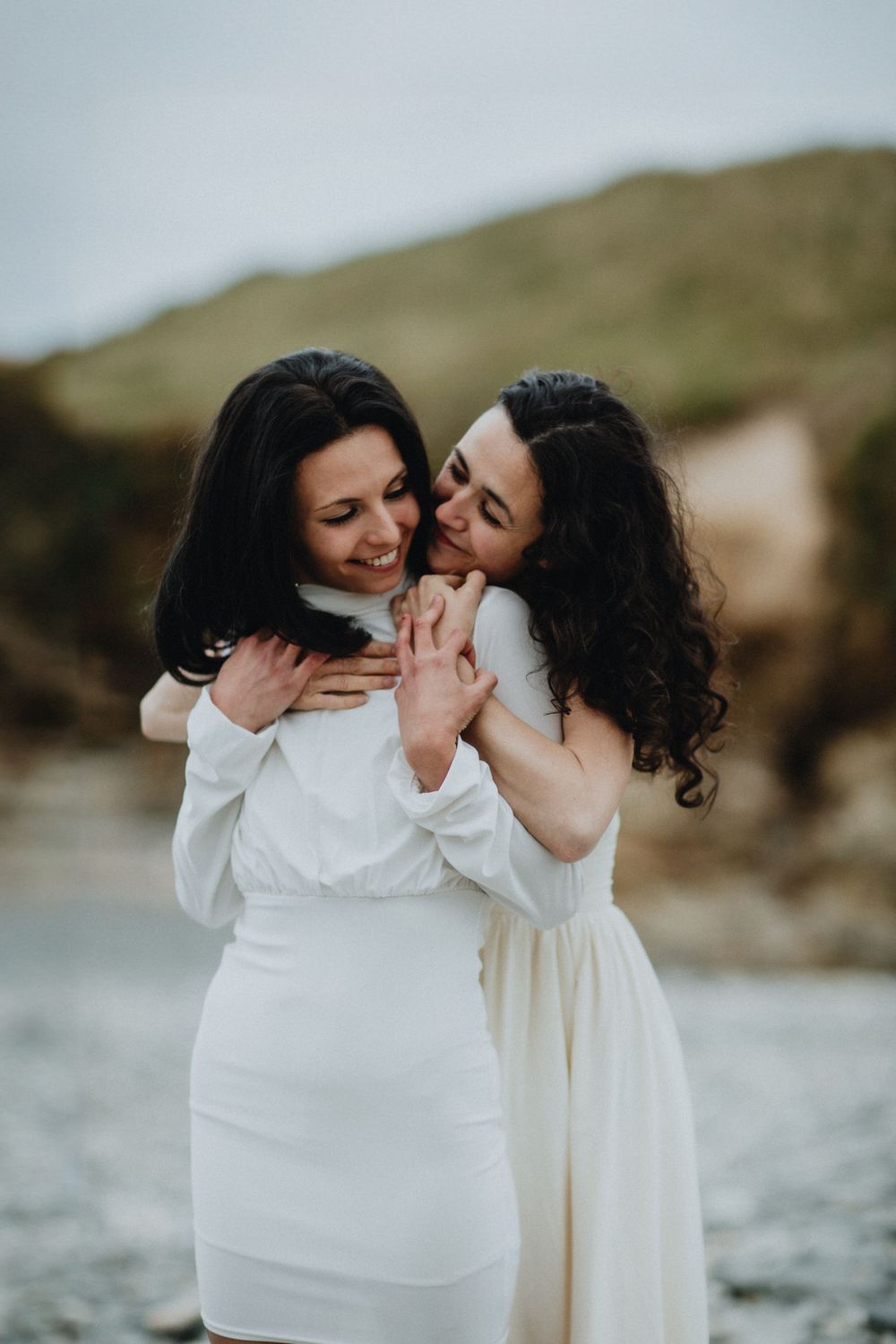 Two people in white dresses share laughter and embrace by the sea.