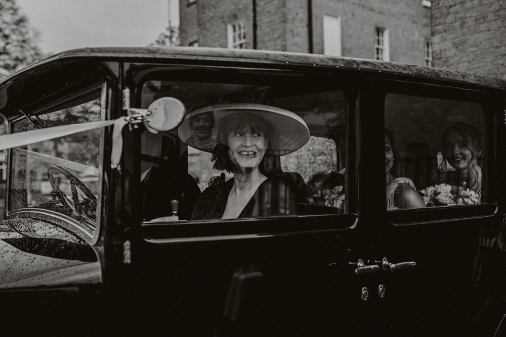 A guest smiles joyfully from inside a vintage black car at a wedding celebration.