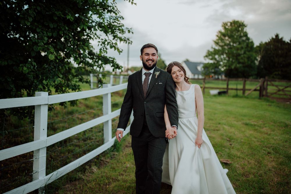 Newlyweds sharing a joyful moment together by a white fence in a countryside setting.
