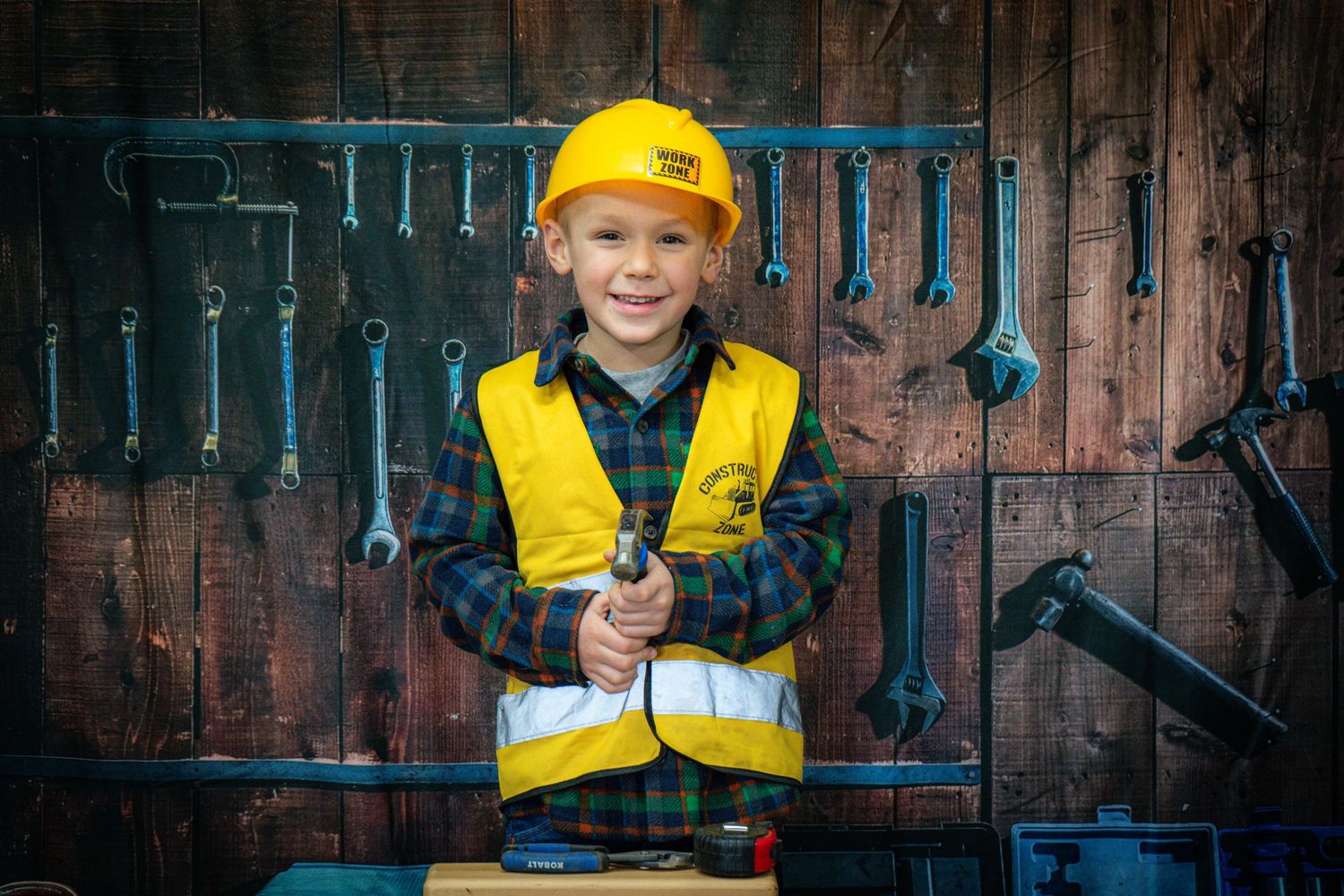 A small construction worker in a yellow safety vest and hard hat holds tools against a wooden wall with hanging equipment.