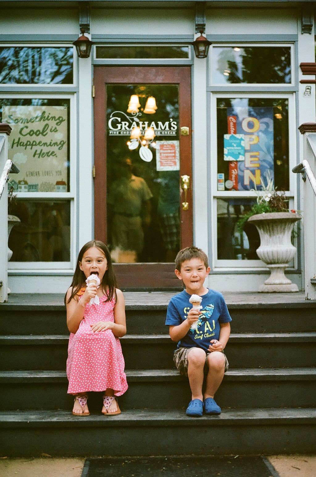 Two smiling children sit on stone steps eating ice cream cones outside Graham's, a storefront with an open sign and a stone planter.