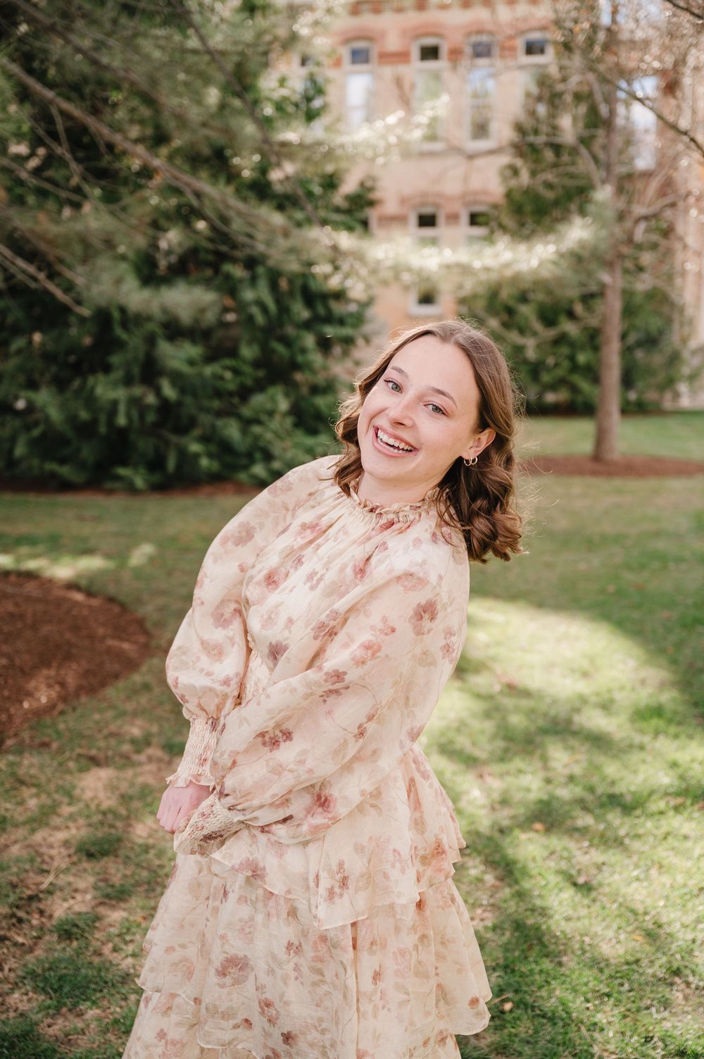 A blush-colored floral dress sways in an outdoor garden setting with a brick building visible in the background.