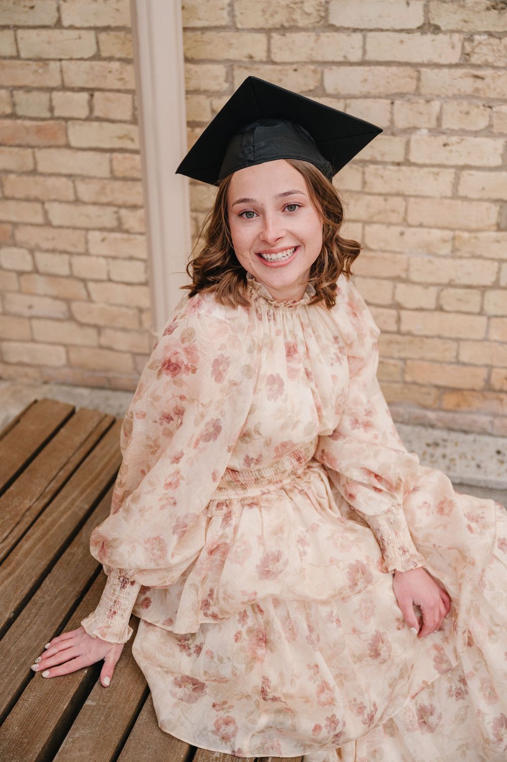 Graduate wearing a black mortarboard cap and floral pink dress sits on wooden bench against brick wall.