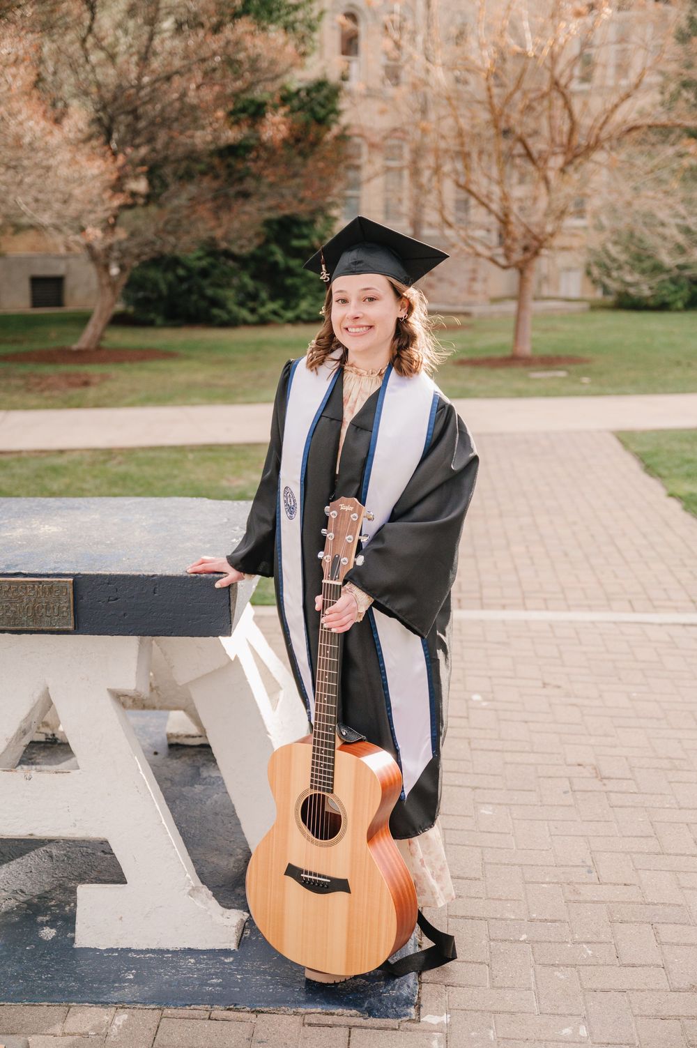 Graduate in cap and gown stands on campus pathway holding an acoustic guitar and diploma on sunny day.