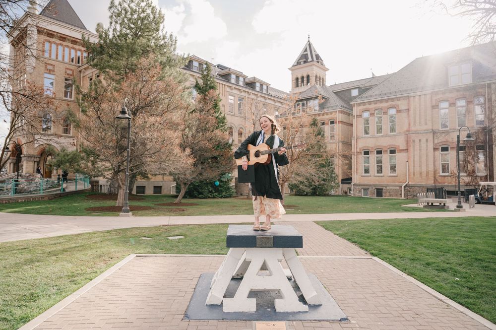 A student jumps joyfully on a giant metal 'A' sculpture on a college campus with historic buildings in the background.