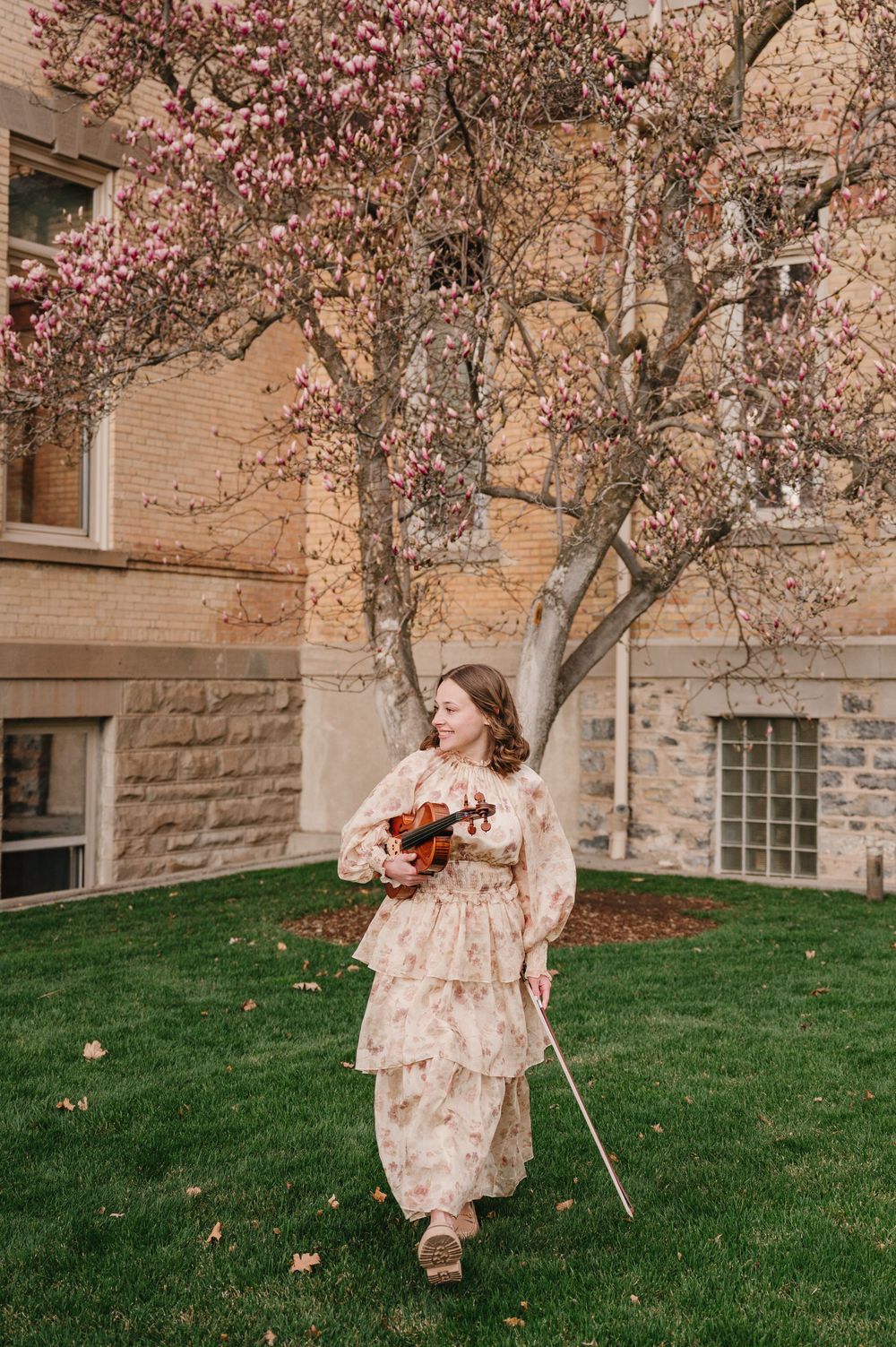 A person in vintage dress stands under a blooming cherry blossom tree holding a violin on a grassy lawn beside a stone building.