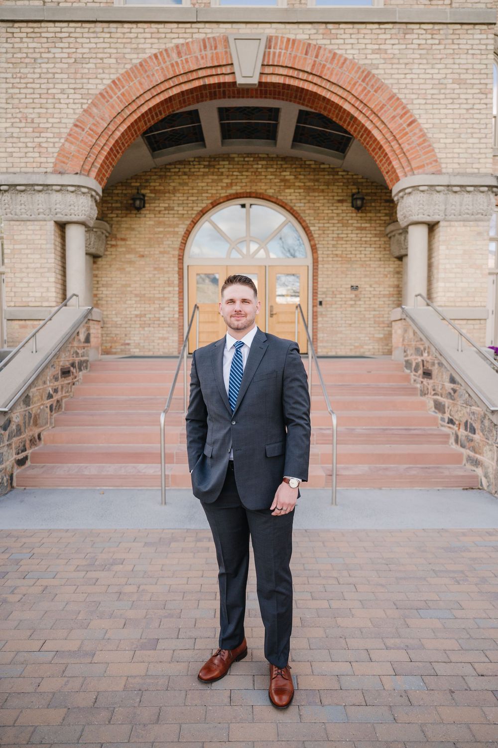 Professional in a dark suit and blue tie stands in front of a brick building entrance with an arched doorway.