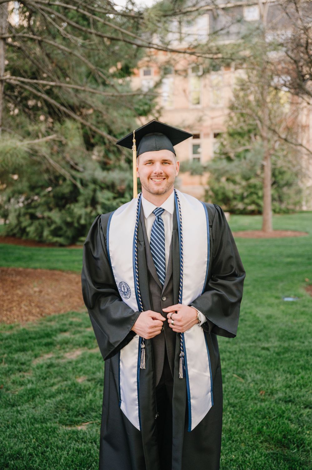 Graduate wearing traditional black graduation gown with white stole and striped tie stands on university lawn.
