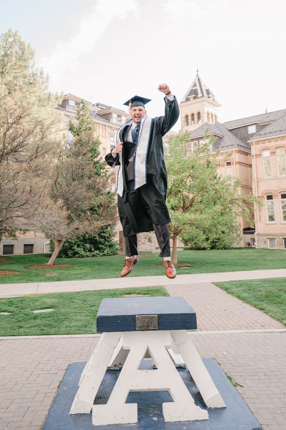 A graduate in black cap and gown jumps joyfully over a large 'A' symbol on a university campus walkway.