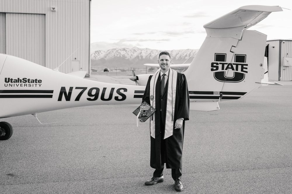 Black and white photo of a person in graduation attire standing next to a State University aircraft on an airfield.