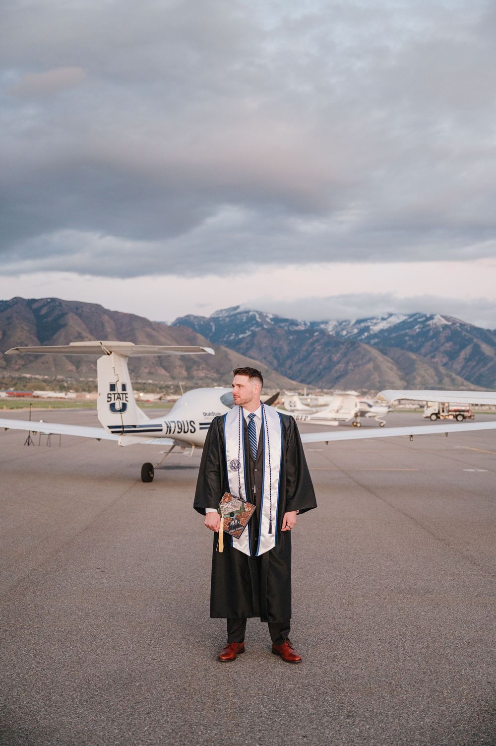 Graduate in cap and gown stands on airport tarmac with small aircraft and snow-capped mountains in background.