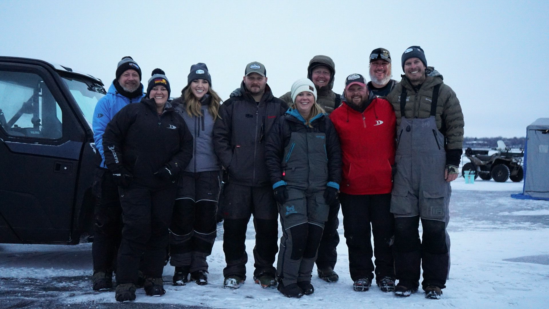 Group gathered on frozen lake wearing winter gear for ice fishing.