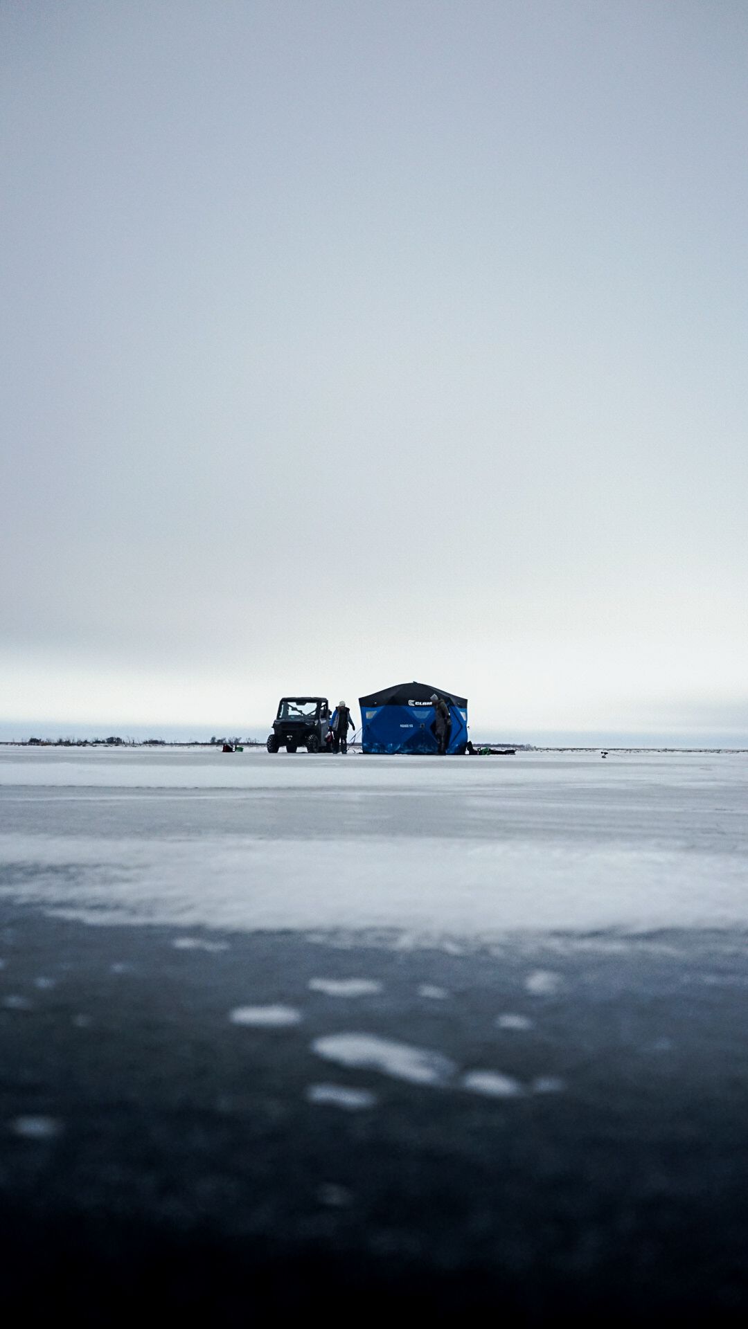 Blue ice fishing shelter on frozen lake with distant figures in a minimalist winter landscape.