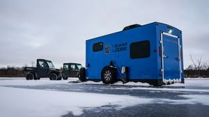 Blue ice fishing shelters and trucks parked on a frozen lake during winter.
