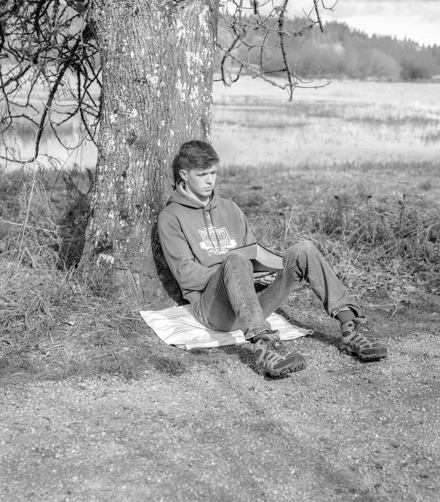 Person relaxing under a tree while reading outdoors in black and white photo.