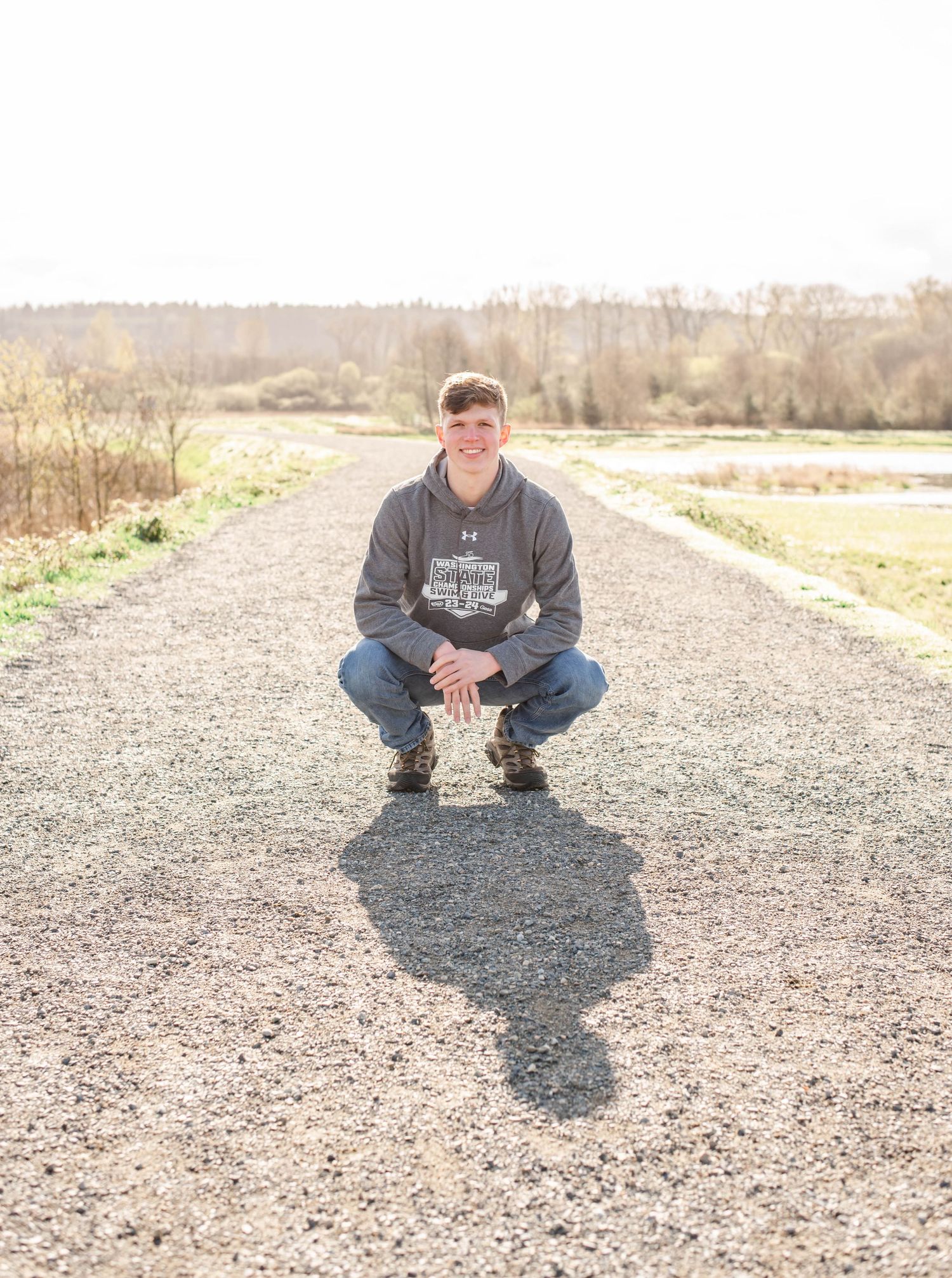 Person crouching on a gravel path with trees and fields in the background.